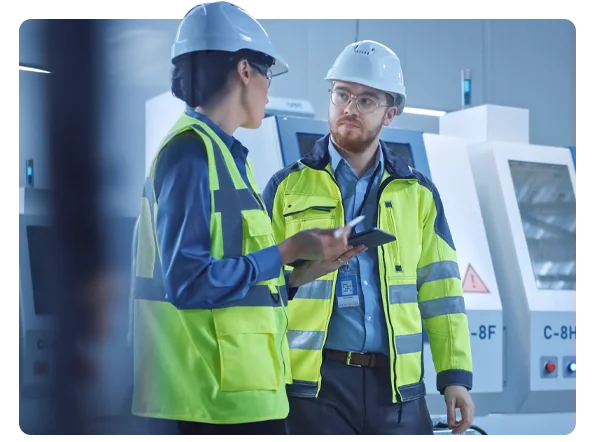 Two engineers in safety helmets and high-visibility jackets discussing production details using a tablet on a modern factory floor.