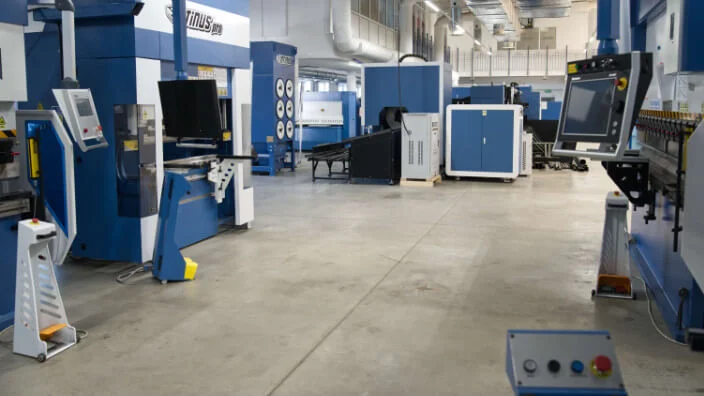 Modern industrial production hall equipped with Otinus CNC machines, featuring press brakes and auxiliary units arranged along a wide concrete aisle with control panels and monitors visible.