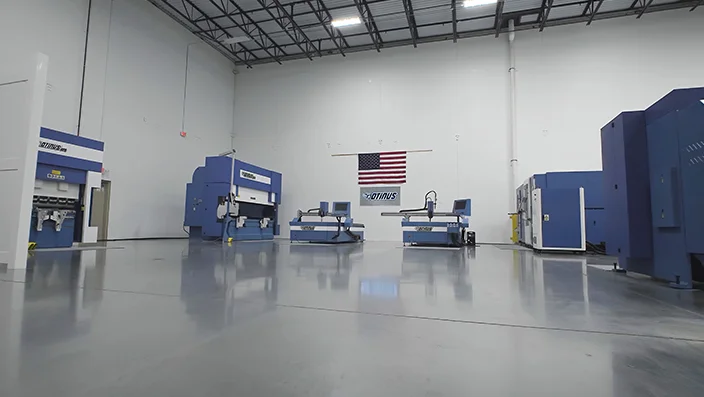Spacious industrial hall with Otinus CNC machines neatly arranged on a polished concrete floor, featuring a large American flag on the back wall and a clean, professional manufacturing setup.